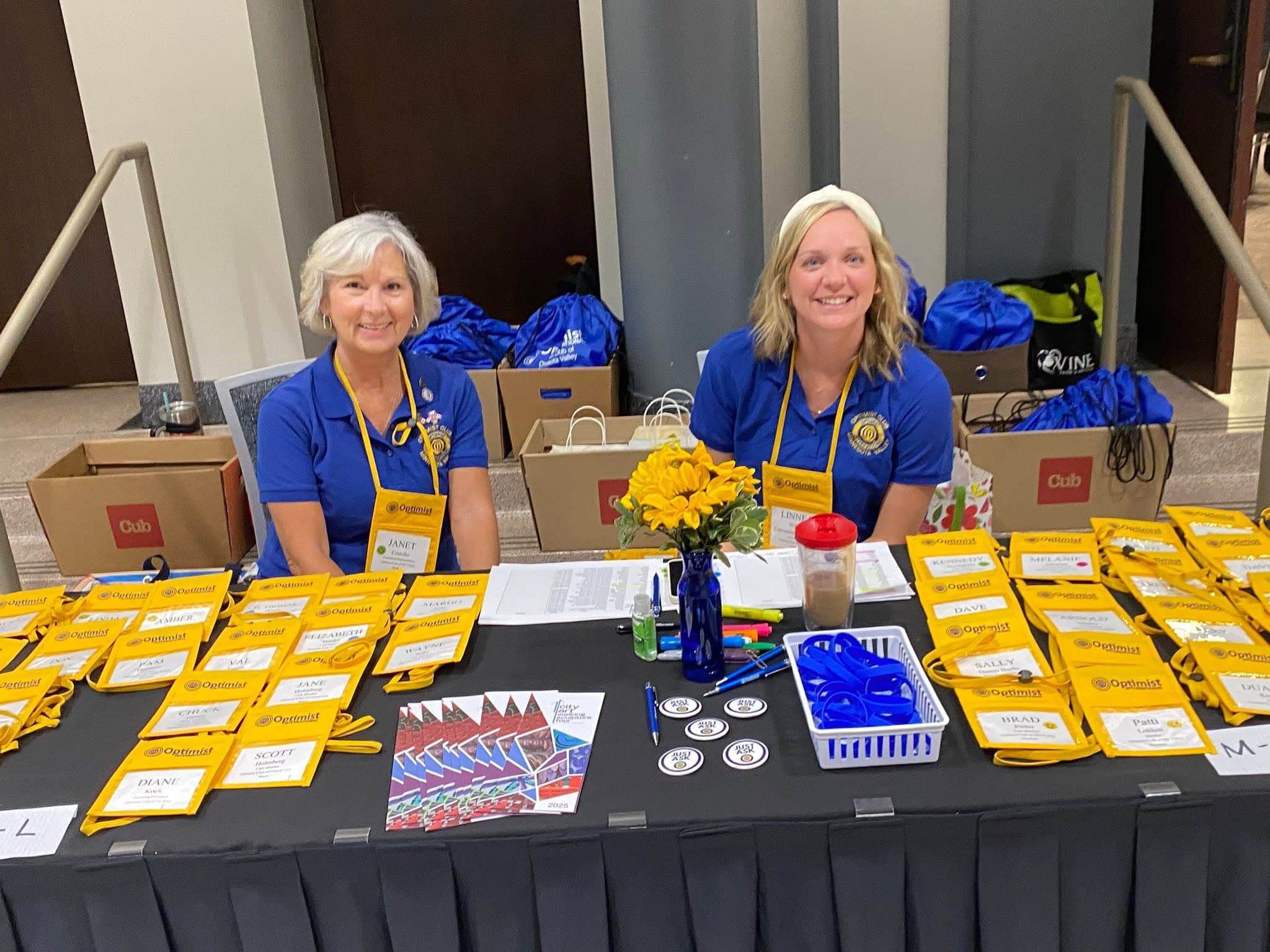 Two Optimist members in club polos at a welcome table covered in name badges and lanyards